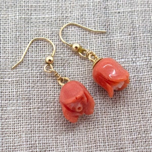 May include: A pair of gold earrings with coral flower charms. The flowers are a light pink colour and have a delicate, detailed design.