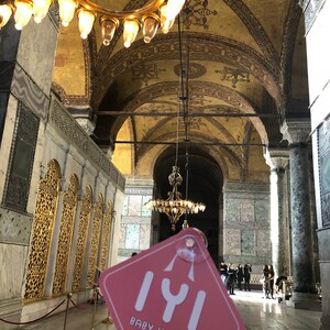 Puede incluir: Un cartel de coche rosa con el texto "Iyi Baby Hatun On Board" en fuente blanca. El cartel est&aacute; colgado de una cuerda y est&aacute; delante de una gran l&aacute;mpara de ara&ntilde;a dorada adornada en una catedral.