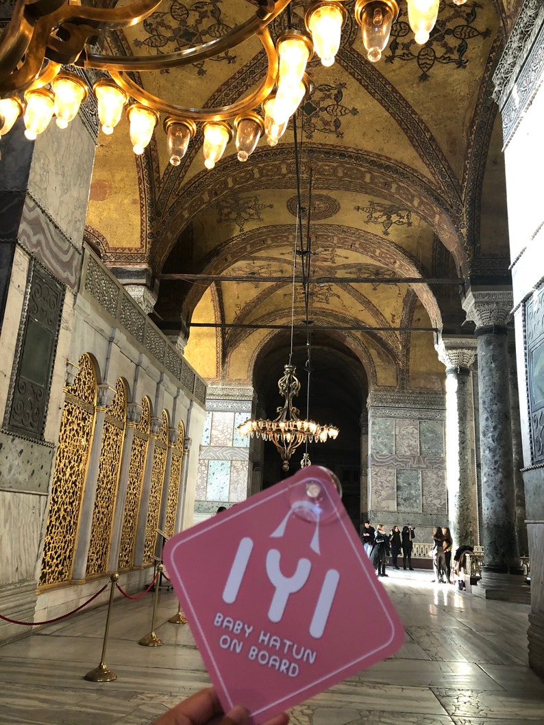 Puede incluir: Un cartel de coche rosa con el texto "Iyi Baby Hatun On Board" en fuente blanca. El cartel est&aacute; colgado de una cuerda y est&aacute; delante de una gran l&aacute;mpara de ara&ntilde;a dorada adornada en una catedral.