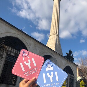 Puede incluir: Dos se&ntilde;ales cuadradas de coche azules y rosas con la palabra turca "Iyi" y el texto "Baby Hatun on Board" y "Baby Alp on Board". Las se&ntilde;ales se sostienen frente a una mezquita con un alto minarete.