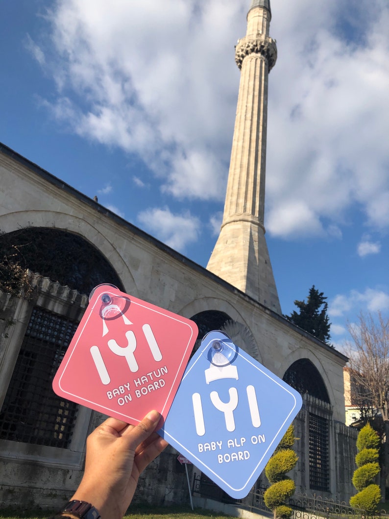 Puede incluir: Dos se&ntilde;ales cuadradas de coche azules y rosas con la palabra turca "Iyi" y el texto "Baby Hatun on Board" y "Baby Alp on Board". Las se&ntilde;ales se sostienen frente a una mezquita con un alto minarete.