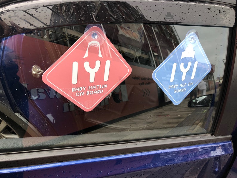 Puede incluir: Dos se&ntilde;ales para la ventana del coche con un dise&ntilde;o de aleta de tibur&oacute;n y el texto "IYI" en blanco. La se&ntilde;al de la izquierda es rosa y dice "Baby Hatun on Board". La se&ntilde;al de la derecha es azul y dice "Baby Alp on Board".