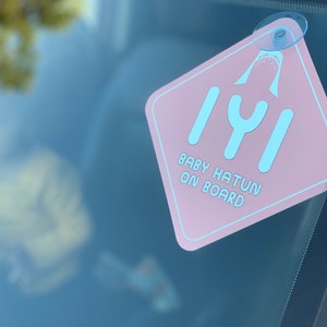 Puede incluir: Un letrero rosa para el coche con una aleta de tibur&oacute;n y el texto "Baby Hatun On Board" en letras azules. El letrero est&aacute; sujeto a la ventana del coche con una ventosa.