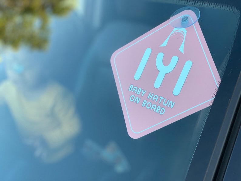 Puede incluir: Un letrero rosa para el coche con una aleta de tibur&oacute;n y el texto "Baby Hatun On Board" en letras azules. El letrero est&aacute; sujeto a la ventana del coche con una ventosa.
