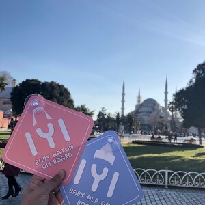 Puede incluir: Dos se&ntilde;ales de coche con un mensaje de beb&eacute; a bordo. La se&ntilde;al de la izquierda es rosa con el texto "Baby Hatun On Board" y la se&ntilde;al de la derecha es azul con el texto "Baby Alp On Board".