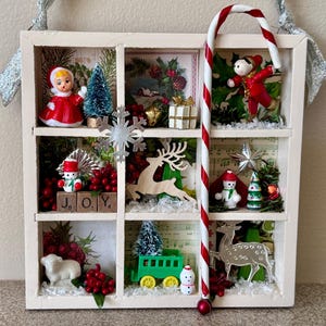 May include: A white framed Christmas shadow box with nine compartments, each containing miniature holiday decorations. Includes a candy cane, snowmen, a reindeer, a doll, a train, and the word "JOY". Silver ribbon hangs from the top.