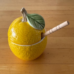 May include: A ceramic lemon-shaped container with a lid, a green leaf, and a wooden spoon. The container is bright yellow with a textured surface. The spoon is inserted into the container. The background is a wooden surface.