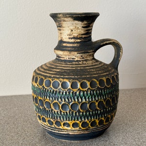 May include: A brown and black ceramic vase with a green and yellow geometric pattern. The vase has a handle and a narrow neck.