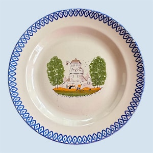 May include: A white ceramic plate with a blue patterned border. The center features a landscape scene with a white building, green trees, and a figure. The plate has a slightly raised edge and a glossy finish.