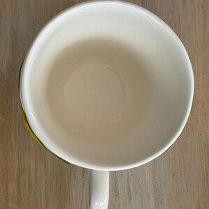 May include: Overhead view of a white ceramic mug with a curved handle. The interior of the mug is a creamy off-white color. The rim of the mug has a thin yellow and green accent. The mug is sitting on a light brown wooden surface.