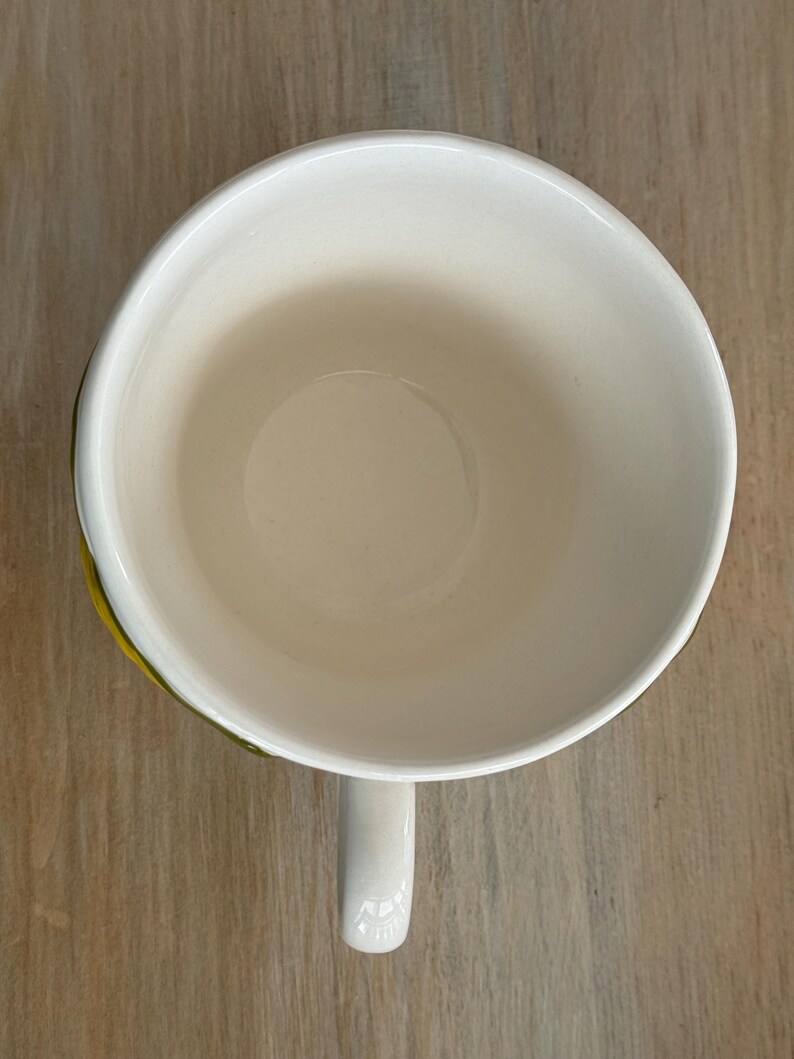 May include: Overhead view of a white ceramic mug with a curved handle. The interior of the mug is a creamy off-white color. The rim of the mug has a thin yellow and green accent. The mug is sitting on a light brown wooden surface.