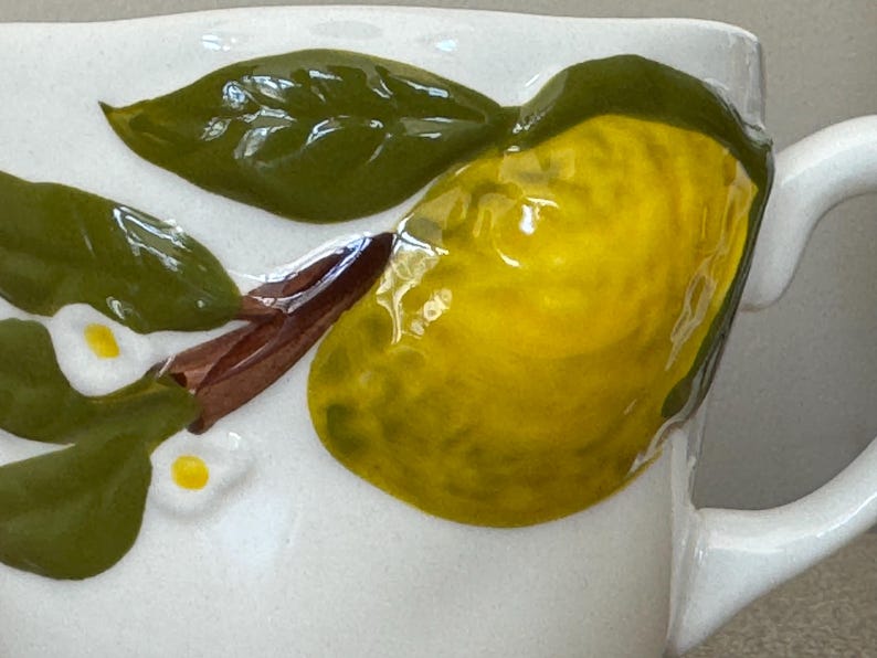 May include: White ceramic mug with a raised lemon and leaf design. The lemon is yellow with green accents, and the leaves are green. The mug has a white handle and a textured surface.