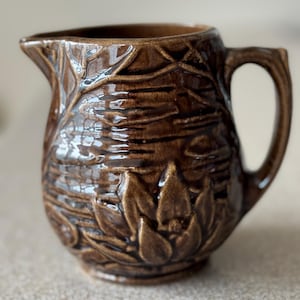 May include: Brown ceramic pitcher with a handle and spout. The pitcher features a raised floral and leaf design. The surface has a glossy finish. The pitcher is likely used for serving beverages.