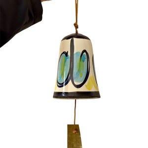 May include: A ceramic wind chime with a cream-colored bell shape, adorned with blue, yellow, and black abstract designs. The bell hangs from a brown cord and has a rectangular brass clapper. The top of the bell is black.