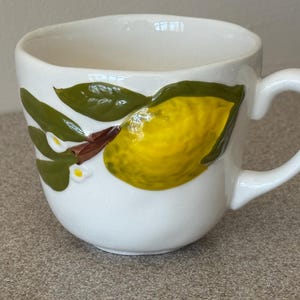 May include: White ceramic mug with a curved handle. The mug features a hand-painted design of a yellow lemon with green leaves and small white flowers. The mug is a kitchenware item.