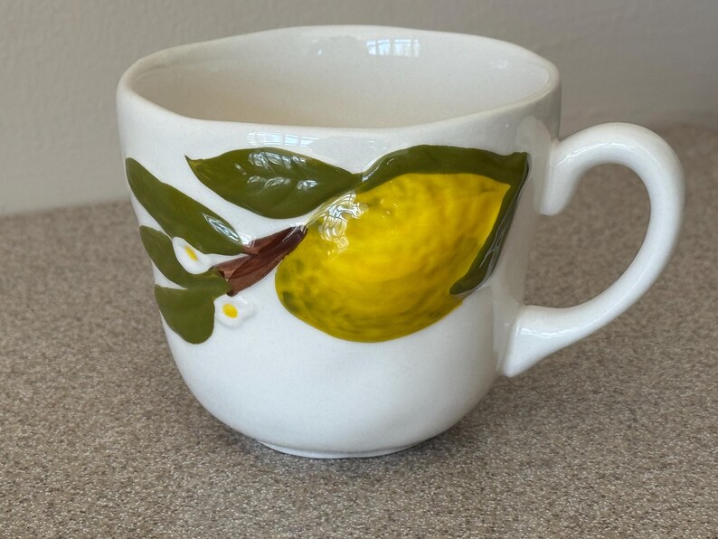 May include: White ceramic mug with a curved handle. The mug features a hand-painted design of a yellow lemon with green leaves and small white flowers. The mug is a kitchenware item.