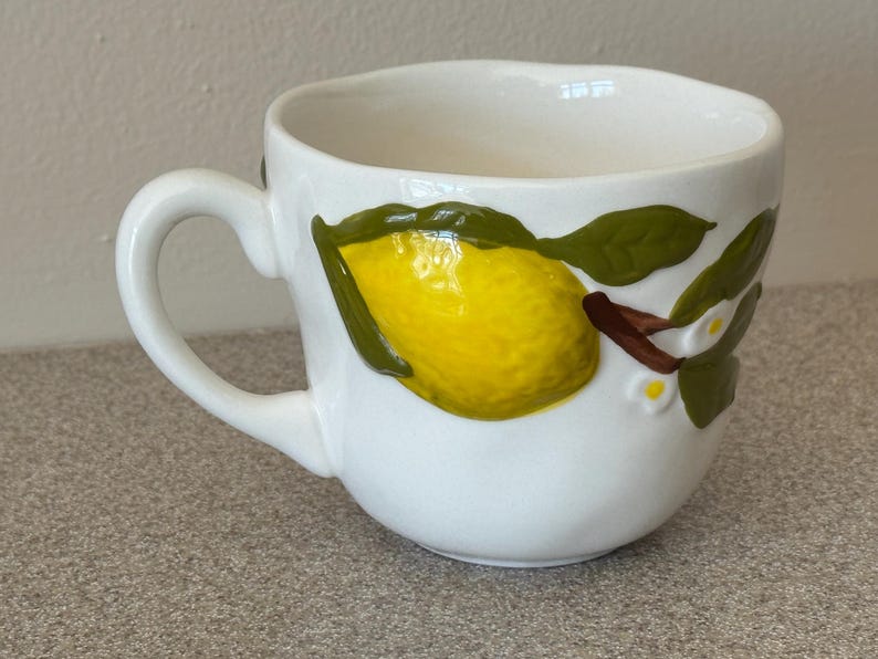 May include: A white ceramic mug with a black handle and the text "But First, Coffee" in a black, handwritten-style font. The mug is on a white surface.