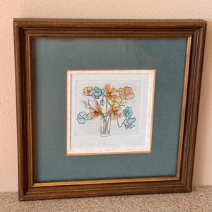 May include: A framed print of a watercolor painting depicting a bouquet of blue and orange flowers in a glass vase. The flowers are stylized and have a whimsical feel. The print is matted and framed in a dark wood frame.