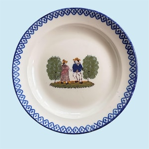 May include: White ceramic plate with a blue geometric border. The center features a colorful illustration of two figures holding hands, standing between two green trees. The plate has a slightly recessed center.
