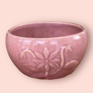 May include: A small, round, pink ceramic bowl with a glossy finish. The bowl features a raised floral design on the exterior. The interior is smooth and the bowl is empty.