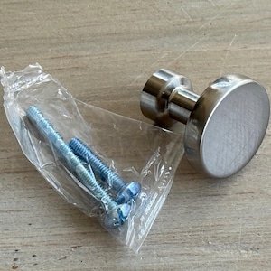 May include: A brushed metal cabinet knob with a flat, circular face and a cylindrical base. Two blue screws are in a clear plastic bag. The hardware is designed for furniture and home improvement projects.