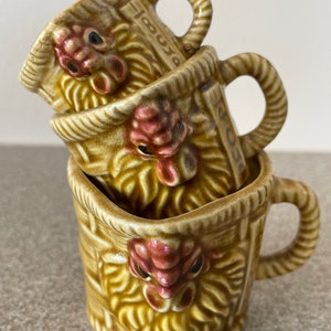 May include: A set of three ceramic measuring cups featuring a rooster design. The cups are brown and yellow with a woven basket pattern and a red rooster face. The cups are stacked on top of each other.