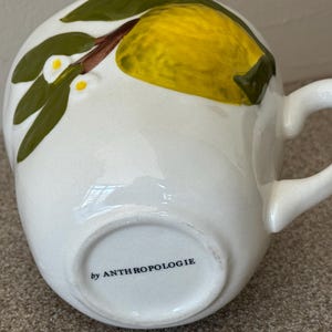 May include: White ceramic mug with a yellow lemon and green leaf design. The mug has a curved handle and the text "by ANTHROPOLOGIE" on the bottom. The mug is a unique piece of kitchenware.