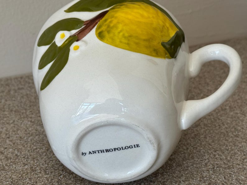 May include: White ceramic mug with a yellow lemon and green leaf design. The mug has a curved handle and the text "by ANTHROPOLOGIE" on the bottom. The mug is a unique piece of kitchenware.