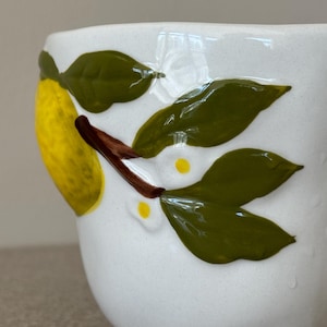 May include: White ceramic mug with a hand-painted lemon and leaf design. The lemon is yellow, and the leaves are green with brown stems. The mug has a slightly textured surface.