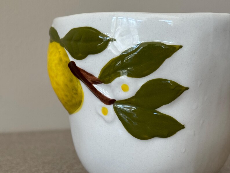 May include: White ceramic mug with a hand-painted lemon and leaf design. The lemon is yellow, and the leaves are green with brown stems. The mug has a slightly textured surface.