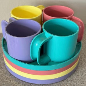 May include: A set of colorful plastic cups and saucers. The cups are in shades of yellow, pink, purple, and teal, with matching saucers in a rainbow of colors. The cups have large, curved handles.