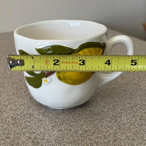 May include: White ceramic teacup with a lemon and leaf design. The cup is approximately 4 inches wide. The handle is on the right side. The cup has a white interior and a white exterior with a lemon and leaf design.