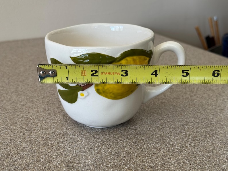 May include: White ceramic teacup with a lemon and leaf design. The cup is approximately 4 inches wide. The handle is on the right side. The cup has a white interior and a white exterior with a lemon and leaf design.