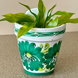 May include: A white ceramic planter with green floral designs and a green plant growing inside.