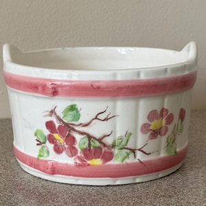 May include: A white ceramic bowl with two small handles, featuring a barrel-like design with horizontal pink bands and a painted floral motif of red flowers with yellow centers and green leaves on the exterior surface of the vessel.