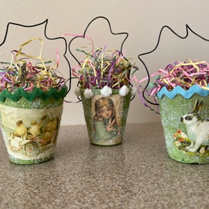 May include: Three decorative Easter buckets with wire handles, filled with colorful paper shreds. Each bucket features a different vintage-style image: chicks in a cart, a girl with a bunny, and a white rabbit. The buckets are green with decorative trim.