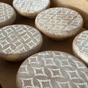 May include: A collection of round, wooden cabinet knobs with a geometric diamond pattern in white. The knobs are a light brown color and appear to be made of wood. The knobs are arranged in a box.