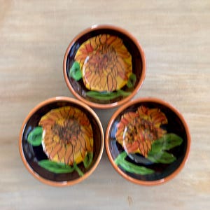 May include: Three round ceramic bowls with a terracotta-colored rim and a dark brown interior. Each bowl features a hand-painted sunflower design in shades of yellow and orange with green leaves, centered at the bottom of the bowl's interior.