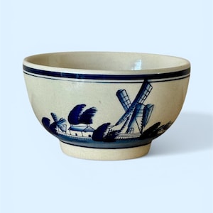 May include: Cream-colored ceramic bowl with a blue windmill design. The bowl features a dark blue band around the rim and a detailed landscape scene with windmills and trees. A decorative piece for serving or display.