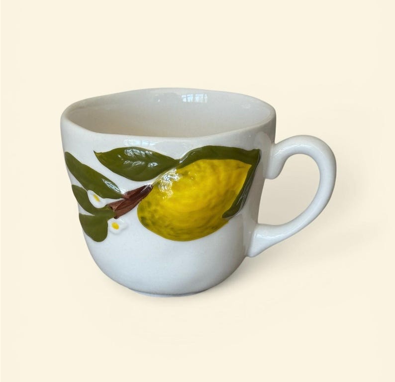 May include: White ceramic mug with a yellow lemon and green leaves design. The mug features a curved handle and a detailed, raised relief of a lemon branch. The design includes a yellow lemon, green leaves, and small white flowers.