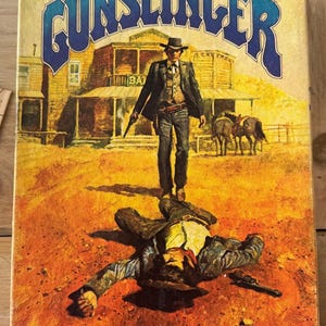 May include: Vintage board game box titled "GUNSLINGER." The cover art depicts a cowboy in a black suit and hat standing over a fallen man. A western town and horses are in the background. The game's title is in blue letters.
