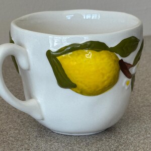 May include: White ceramic mug with a yellow lemon and green leaves design. The mug has a curved handle and a slightly irregular rim. The lemon is textured and appears to be hand-painted, adding a unique touch to the mug.
