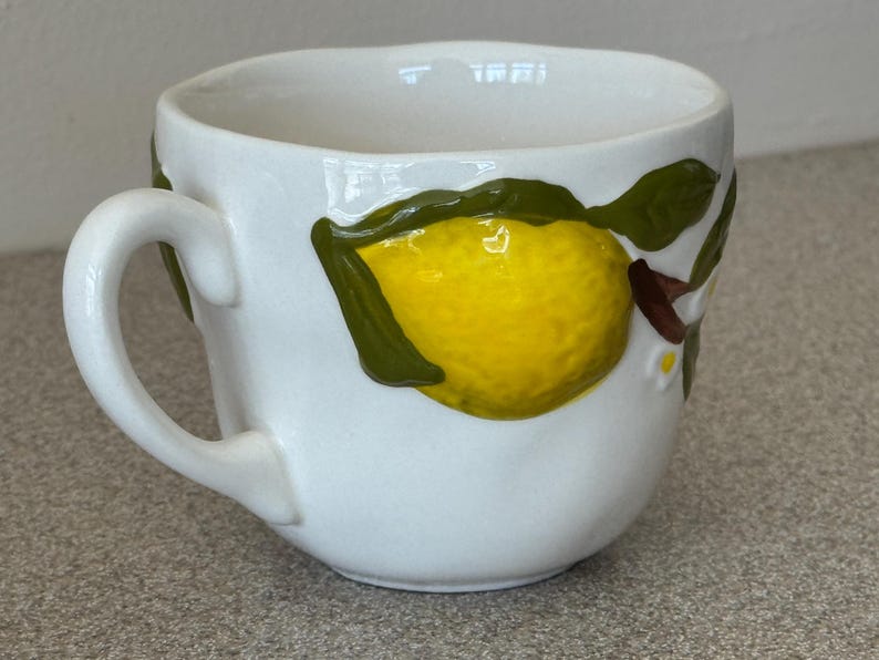 May include: White ceramic mug with a yellow lemon and green leaves design. The mug has a curved handle and a slightly irregular rim. The lemon is textured and appears to be hand-painted, adding a unique touch to the mug.