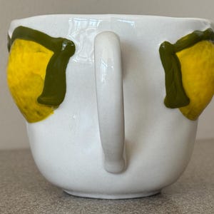 May include: White ceramic mug with a curved handle. The mug is decorated with two yellow lemons with green leaves. The lemons are painted on the front of the mug. The mug is sitting on a gray surface.