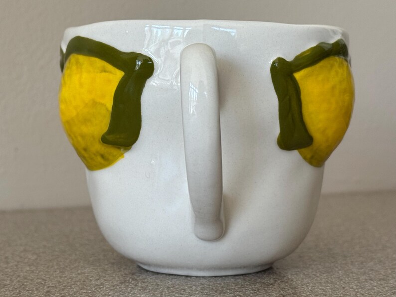May include: White ceramic mug with a curved handle. The mug is decorated with two yellow lemons with green leaves. The lemons are painted on the front of the mug. The mug is sitting on a gray surface.