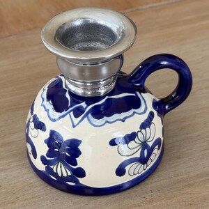 May include: A ceramic candle holder with a rounded base featuring a cream background and blue floral patterns. The piece includes a small side handle and a metallic silver-toned top insert designed to hold a single taper candle securely.