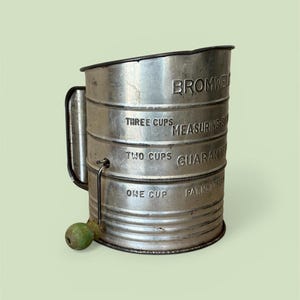 May include: Vintage metal sifter with a handle and a green wooden knob. The sifter has the text "BROMWEL" and "THREE CUPS MEASURING-S" and "TWO CUPS GUARANTEED" and "ONE CUP PAT. No 1753" printed on it. The sifter is a kitchen tool.