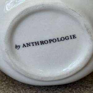 May include: Close-up of the bottom of a white ceramic bowl. The words "by ANTHROPOLOGIE" are printed in black, centered on the base. The bowl has a rounded shape and a smooth, glossy finish.