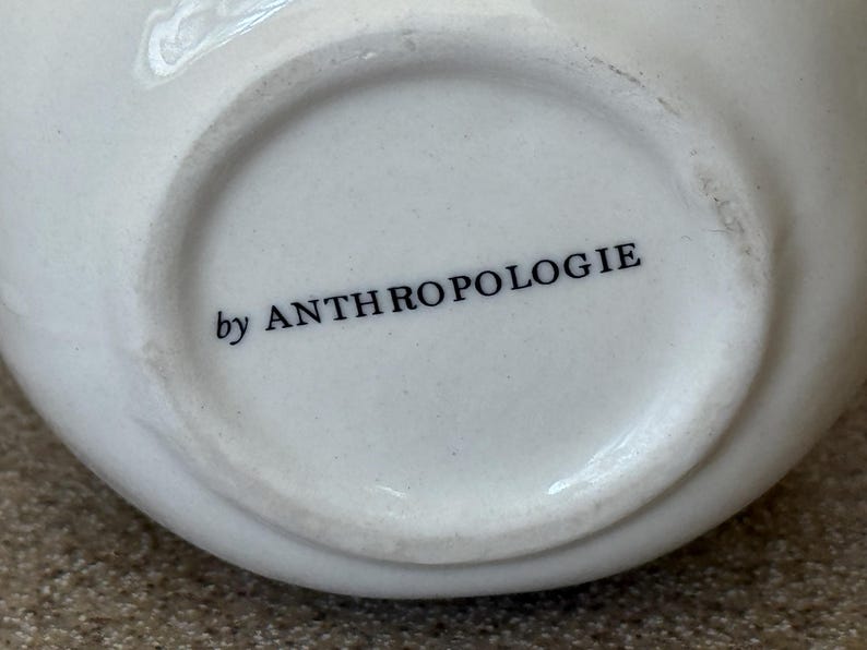 May include: Close-up of the bottom of a white ceramic bowl. The words "by ANTHROPOLOGIE" are printed in black, centered on the base. The bowl has a rounded shape and a smooth, glossy finish.