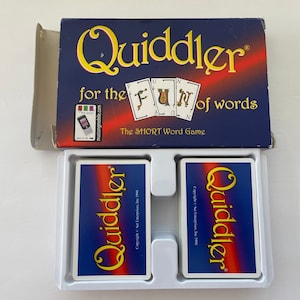 May include: A Quiddler card game box set. The navy blue box features the game title in yellow script and the phrase "for the fun of words." Two decks of cards are visible in a white tray, each with the game title.